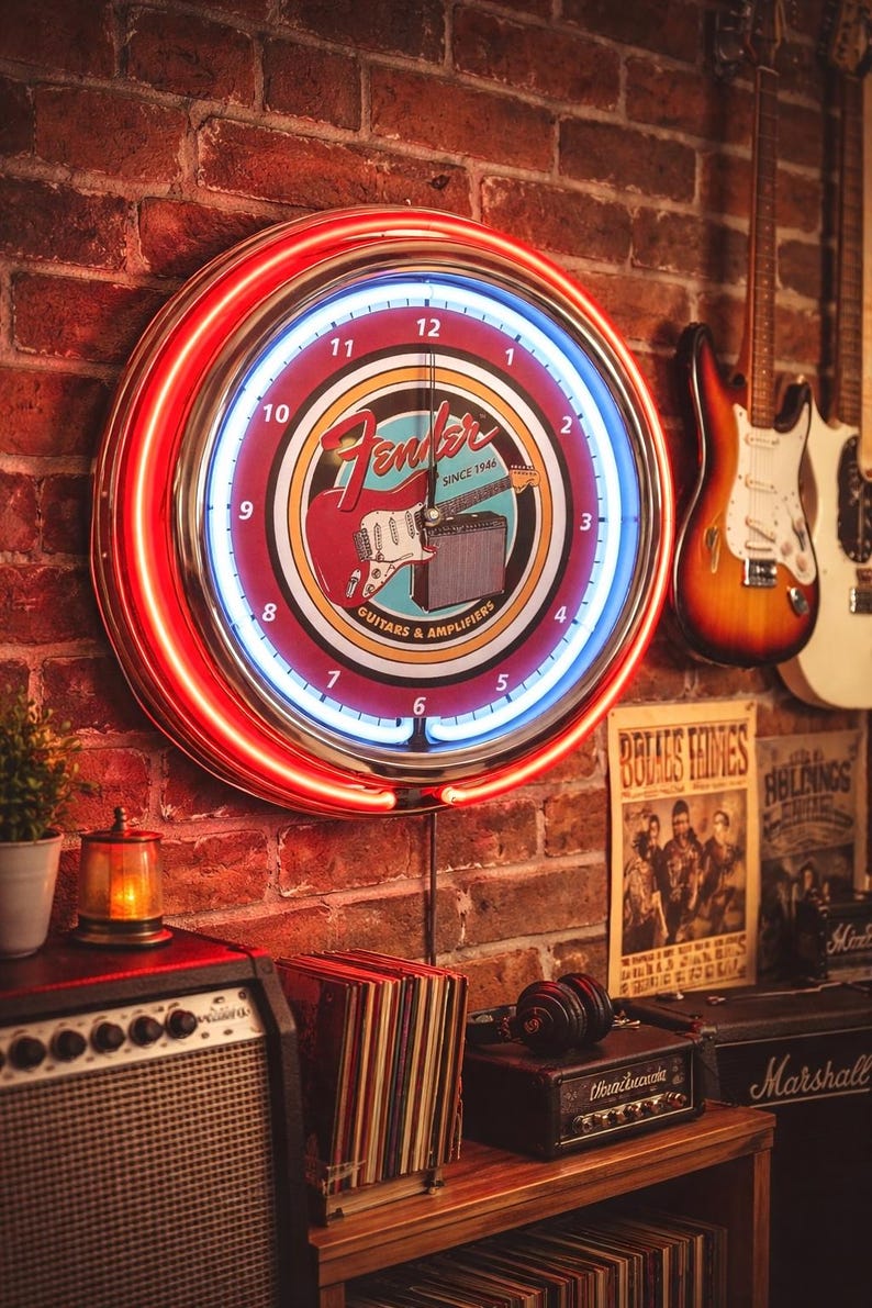 May include: A Fender neon clock with a red and blue illuminated ring, mounted on a brick wall. The clock face features a guitar and amplifier graphic. Guitars and amplifiers are also visible in the background, along with vintage posters and a stereo.