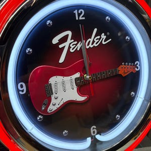 May include: Circular wall clock with a red and black face featuring a red electric guitar graphic. The clock has a white neon light border and the Fender logo. The clock hands are black and red.