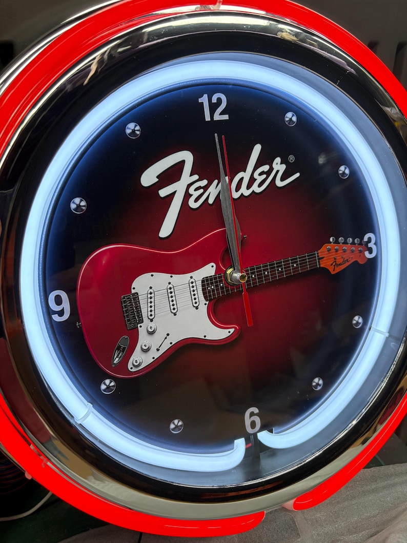 May include: Circular wall clock with a red and black face featuring a red electric guitar graphic. The clock has a white neon light border and the Fender logo. The clock hands are black and red.