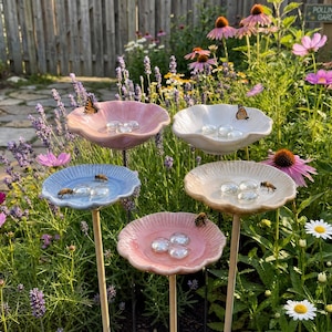 May include: Five ceramic bird baths in pastel shades of pink, blue, and cream, each with a scalloped edge. They are mounted on wooden stakes. Butterflies and bees are visible, set against a backdrop of a vibrant garden filled with flowers.
