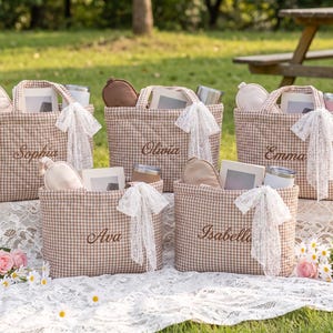 May include: Five quilted tote bags in a brown and white gingham pattern, each embroidered with a unique name: Sophia, Olivia, Emma, Ava, and Isabella. Each bag features a white lace bow, a sleep mask, a small book, and a metal tumbler inside.