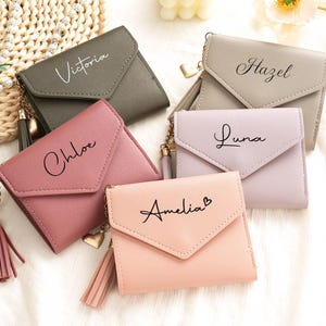 Puede incluir: Cinco carteras pequeñas y personalizadas en tonos rosa, gris y verde están dispuestas sobre una superficie blanca. Cada cartera tiene un nombre impreso en la parte delantera: Victoria, Chloe, Amelia, Hazel y Luna. Las carteras tienen una borla y un dije de corazón.