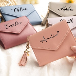 Puede incluir: Carteras personalizadas en colores pastel, como rosa, azul y gris. Cada cartera presenta un nombre en escritura elegante. Las carteras tienen un cierre de solapa y un adorno de borla adjunto a una cadena.