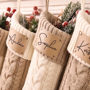 May include: Four knit Christmas stockings in cream and beige tones, each featuring a cable knit pattern, a ribbed cuff, and a personalised tan leather-look patch with a name in black script. Each stocking contains artificial pine and red berries.