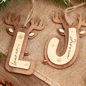 May include: Two wooden Christmas ornaments shaped as reindeer, with the letters "L" and "J" cut out. The letter "L" has the name "Lauren" and the letter "J" has the name "Jessica" written on them. Each ornament has a snowflake design and a red nose.