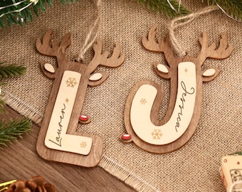 Puede incluir: Dos adornos navideños de madera con forma de reno, con las letras "L" y "J" recortadas. La letra "L" tiene el nombre "Lauren" y la letra "J" tiene el nombre "Jessica" escritos. Cada adorno tiene un diseño de copo de nieve y una nariz roja.