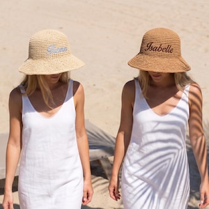 May include: Two woven straw bucket hats, one light beige with the name Fiona embroidered in white, and one tan with the name Isabelle embroidered in dark brown. Both hats feature a rounded crown and a downward-sloping brim, worn with white dresses.