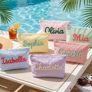 May include: A collection of six rectangular zipped pouches made of seersucker fabric in various pastel colours, each featuring a name embroidered in a contrasting colour: Isabella, Sophia, Olivia, Mia, Camelia, and Charlotte.