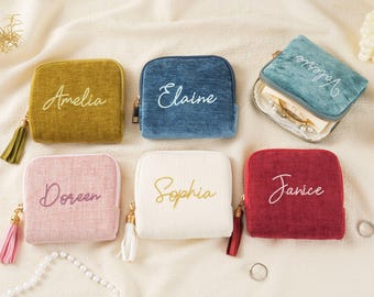 Könnte beinhalten: Eine Auswahl an kleinen, quadratischen Reißverschlusstaschen in verschiedenen Farben: Olivgrün, Blau, Rosa, Weiß und Rot. Jede Tasche ist mit einem Namen bestickt und hat eine Quaste am Reißverschluss.