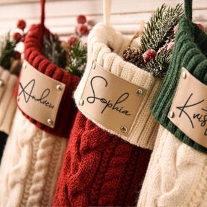 Op de afbeelding: Een rij van vier kerstsokken van kabelgebreid materiaal in de kleuren rood, groen en crème. Elke sok is voorzien van een beige label in lederlook, bevestigd met zilveren klinknagels, met de namen George, Andrew, Sophia en Kristin in zwarte letters.