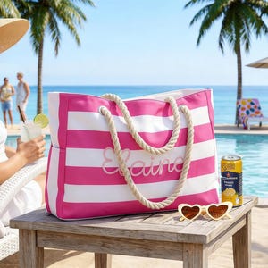 Puede incluir: Un bolso tote a rayas rosas y blancas con asas de cuerda y el nombre "Elaine" bordado en la parte delantera. La bolsa está sobre una mesa de madera con gafas de sol en forma de corazón y una lata de bebida. Una mujer con sombrero para el sol está cerca.
