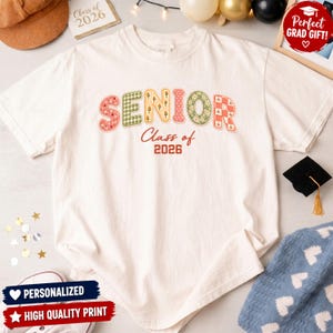 Pode incluir: Camiseta creme com a palavra "SENIOR" em letras de patchwork coloridas e "Class of 2026" impressa abaixo. Esta peça de vestuário com tema de formatura é perfeita para celebrar a turma de 2026.