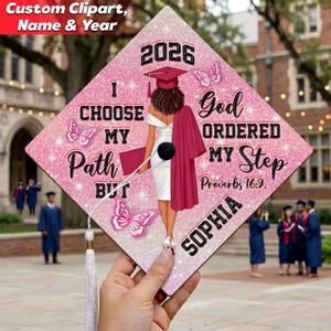 May include: A pink graduation cap with a glittery background, featuring a graphic of a graduate in a white dress and maroon graduation gown. The cap includes the year 2026, butterflies, and the text "I Choose My Path But God Ordered My Step."