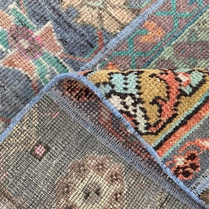May include: Close-up of the edge of a blue, green, orange, and brown patterned rug with a blue fringe. The rug is woven with a variety of textures and colors.