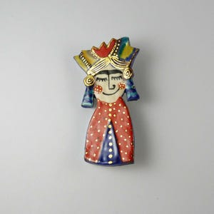 Può includere: Una spilla in ceramica a forma di figura stilizzata che indossa un copricapo con accenti dorati. La figura ha un viso color crema con lineamenti neri, guance rosse e un vestito rosso a pois bianchi con un pannello triangolare blu.