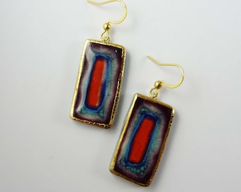 May include: A pair of rectangular earrings with gold-colored hooks and frames. The earrings feature a central red rectangle surrounded by blue and purple hues, with a gold border. The design is abstract and artistic.