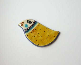 May include: A yellow ceramic bird with black polka dots, a blue and white eye, and gold accents. The bird has a blue and white stripe around its neck.