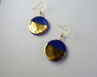 May include: A pair of drop earrings featuring circular blue discs. Each disc is divided horizontally, with the bottom half finished in a reflective gold metallic tone and the top half in a solid deep blue. The earrings hang from gold-toned hooks.