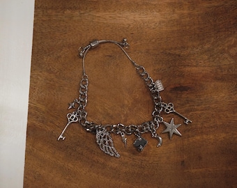May include: A silver charm necklace with a variety of charms, including keys, a star, a wing, and a small box. The necklace is on a wooden surface.