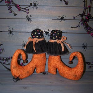 May include: A pair of orange witch's boots with black spiderweb patterns and black ruffles. The boots are on a wooden surface with a black vine and purple berries, and black spiders.