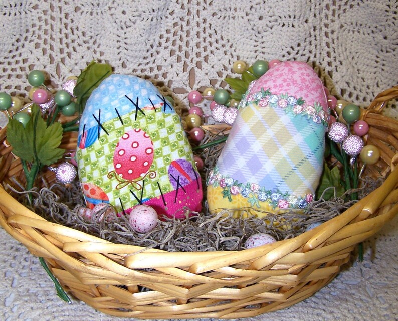 K&ouml;nnte beinhalten: Zwei handgefertigte Stoff-Ostereier in einem Weidenkorb. Ein Ei ist blau, gr&uuml;n und rosa mit einem karierten Muster. Das andere Ei ist gelb, rosa und lila mit einem karierten Muster. Beide Eier haben gestickte Details.