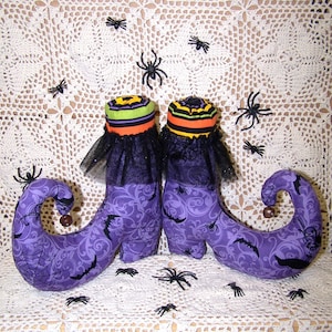 May include: A pair of purple witch's boots with black bats and spiderwebs printed on them. The boots have a pointed toe and a curled up heel. The boots are decorated with black lace and a green and orange striped cuff. The boots are sitting on a white crocheted surface with black spiders scattered around them.
