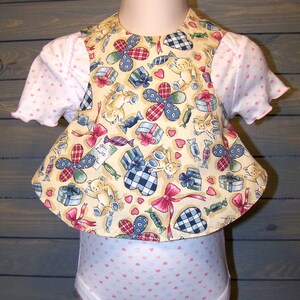 May include: A yellow, white, blue, and red fabric baby bib with a teddy bear, candy, and gift box pattern. The bib is attached to a white bodysuit with a pink polka dot pattern.
