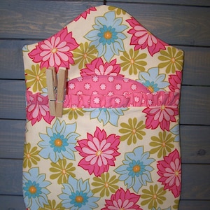 May include: A fabric organizer with a floral pattern in pink, blue, and green on a cream background. The organizer has a pocket with a ruffled edge and wooden clothespins attached. It hangs from a metal hanger.