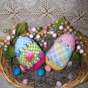 Könnte beinhalten: Zwei handgefertigte Stoff-Ostereier in einem Weidenkorb mit grünen und rosa floralen Akzenten. Ein Ei ist rosa und weiß kariert, das andere ist grün und rosa Patchwork mit den Buchstaben 'XIX' darauf gestickt.