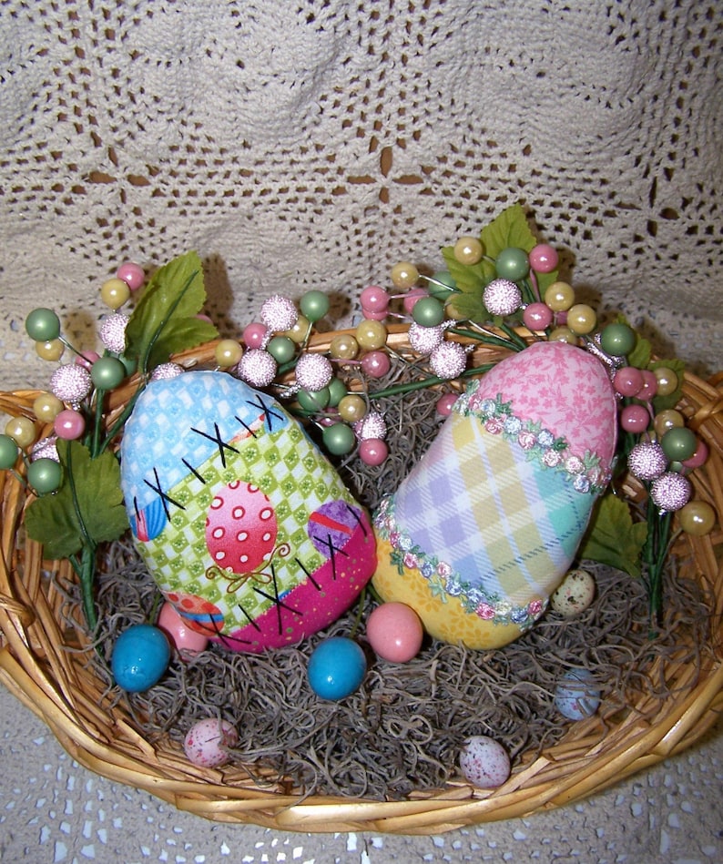 K&ouml;nnte beinhalten: Zwei handgefertigte Stoff-Ostereier in einem Weidenkorb mit gr&uuml;nen und rosa floralen Akzenten. Ein Ei ist rosa und wei&szlig; kariert, das andere ist gr&uuml;n und rosa Patchwork mit den Buchstaben 'XIX' darauf gestickt.