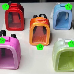 May include: Five colorful miniature backpacks in red, orange, blue, pink, and green. Each backpack has a clear front panel and black straps. The backpacks are arranged on a white surface.