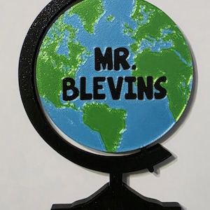 May include: A globe-shaped decoration with a blue and green world map design. The text "MR. BLEVINS" is displayed in black letters on the globe. The globe is supported by a black stand.