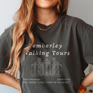 May include: A dark gray t-shirt with a white graphic design that says 'Pemberley Walking Tours' with a drawing of a house and the text 'Hertfordshire Derbyshire For those 'very fond of walking'.