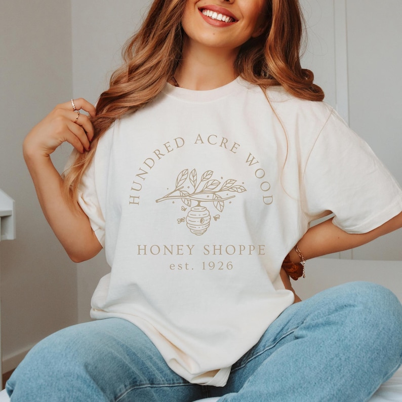 May include: A white t-shirt with a brown logo that says 'Hundred Acre Wood Honey Shoppe est. 1926' with a drawing of a beehive on a branch with leaves.