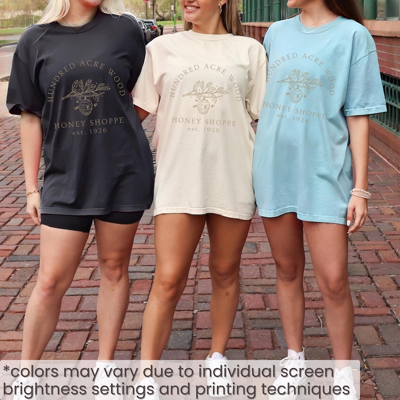 May include: Three t-shirts in charcoal, cream, and light blue, each with a gold graphic and the text "HUNDRED ACRE WOOD HONEY SHOPPE est. 1926." The shirts are oversized and worn with shorts and sneakers. The image text says "colors may vary due to individual screen brightness settings and printing techniques."