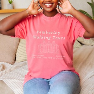 Op de afbeelding: Koraalrood T-shirt met witte tekst "Pemberley Walking Tours" en een architectonische tekening. Het shirt bevat ook de teksten "Hertfordshire" en "Derbyshire" met de zin "For those very fond of walking."