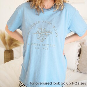 May include: Light blue t-shirt with a graphic design in the center. The design includes the words "Hundred Acre Wood" in a circular pattern, with "Honey Shoppe est. 1926" below a bee-themed illustration. The text "for oversized look go up 1-2 sizes" is at the bottom.