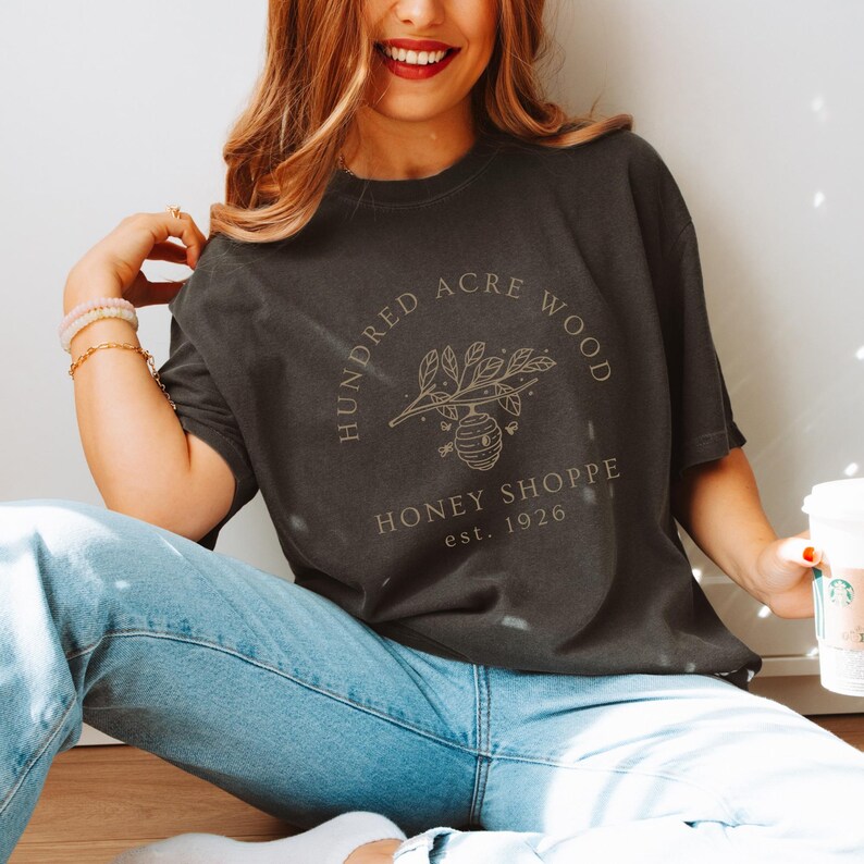 May include: Dark gray t-shirt with a circular graphic that reads "Hundred Acre Wood Honey Shoppe est. 1926". The design features a honeybee hive and sprigs of leaves. The shirt is worn with blue jeans.