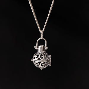 Puede incluir: Un collar con colgante plateado con un dije redondo de estilo filigrana. El dije tiene un diseño calado y adornado y cuelga de una delicada cadena. El fondo es negro liso.