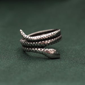 Puede incluir: Un anillo de metal plateado con forma de serpiente enroscada. La banda presenta un patrón de escamas detallado y texturizado que se envuelve alrededor del dedo, con la cabeza de la serpiente en un extremo y la cola afilada en el otro extremo.