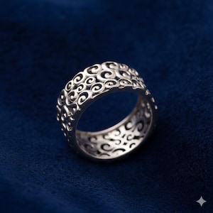 Puede incluir: Un anillo de plata con un diseño calado de patrones arremolinados. El anillo se encuentra sobre un fondo azul oscuro, resaltando sus intrincados detalles y brillo metálico. El diseño del anillo es consistente alrededor de toda la banda.