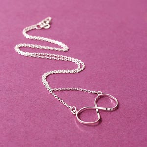 Könnte beinhalten: Eine silberfarbene Halskette mit einem Anhänger in Form eines Unendlichkeitszeichens. Kette und Anhänger bestehen aus glänzendem Metall, vor einem einfarbigen lila Hintergrund. Das Unendlichkeitszeichen ist eine durchgehende Schleife.