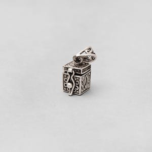 May include: A small, silver-colored pendant in the shape of a box with intricate, openwork designs. The box has a hinged lid and a decorative clasp. The pendant has a loop for attaching to a chain. The background is a solid, light gray.