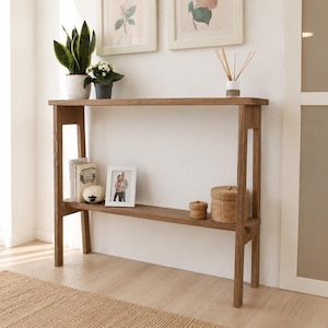Op de afbeelding: Een houten console met twee niveaus in lichtbruin. De tafel heeft een minimalistisch ontwerp met een bovenste plank voor planten en decoratie, en een onderste plank met ingelijste foto's en geweven manden. Ideaal voor gangen of woonkamers.