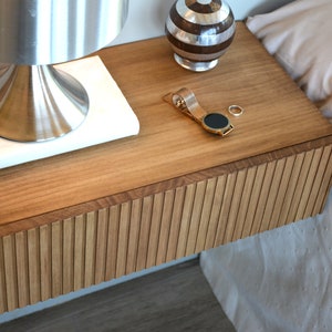 Puede incluir: Una mesita de noche flotante de madera con frente de caj&oacute;n de listones, que incluye una l&aacute;mpara met&aacute;lica sobre un libro blanco, una peque&ntilde;a esfera decorativa a rayas, un reloj de pulsera dorado y un anillo plateado sobre la superficie.
