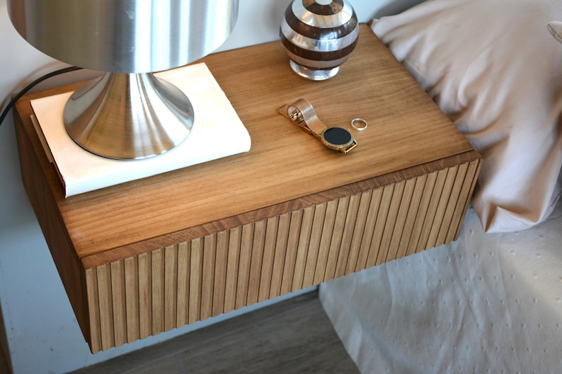 Puede incluir: Una mesita de noche flotante de madera con frente de caj&oacute;n de listones, que incluye una l&aacute;mpara met&aacute;lica sobre un libro blanco, una peque&ntilde;a esfera decorativa a rayas, un reloj de pulsera dorado y un anillo plateado sobre la superficie.