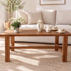 May include: A rectangular wooden coffee table with four sturdy legs and a lower support crossbar. The table surface is a warm brown wood grain, holding a white ceramic teapot, a matching sugar bowl, two teacups on saucers, and a small potted plant.