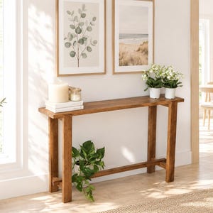 Peut inclure: Une console rectangulaire en bois avec une finition brun moyen, dotée de quatre pieds droits et d'une traverse inférieure. Le plateau supporte deux bougies blanches sur des livres et deux petits pots blancs avec des plantes vertes.