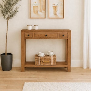 Peut inclure: Une console rectangulaire en bois dotée de trois tiroirs avec de petits boutons carrés noirs. La table possède une étagère inférieure contenant un panier en osier tressé avec deux tasses et soucoupes blanches, et deux bocaux blancs sur le dessus.