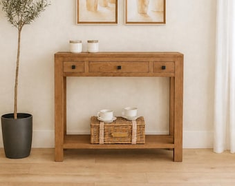 May include: A rectangular wooden console table featuring three drawers with small black square knobs. The table has a lower shelf holding a woven wicker basket with two white ceramic cups and saucers placed on top of it, and two white jars on the top surface.